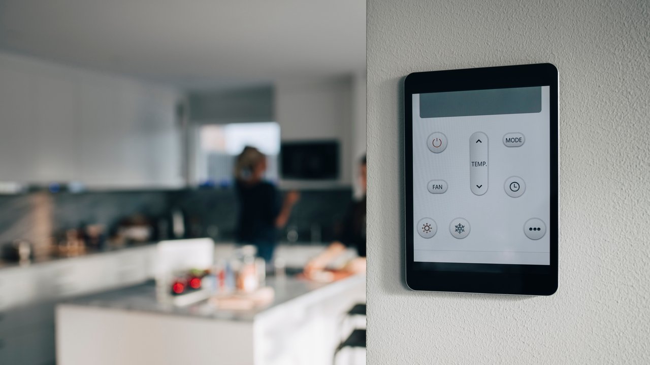 A digital control panel mounted on a wall in a kitchen, with blurred figures in the background.