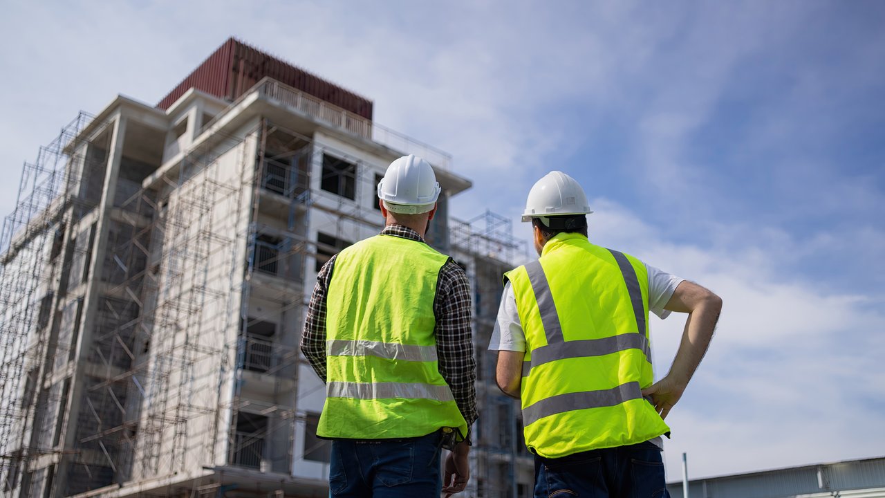 Two construction workers in safety vests and helmets observe a building under construction with scaffolding.