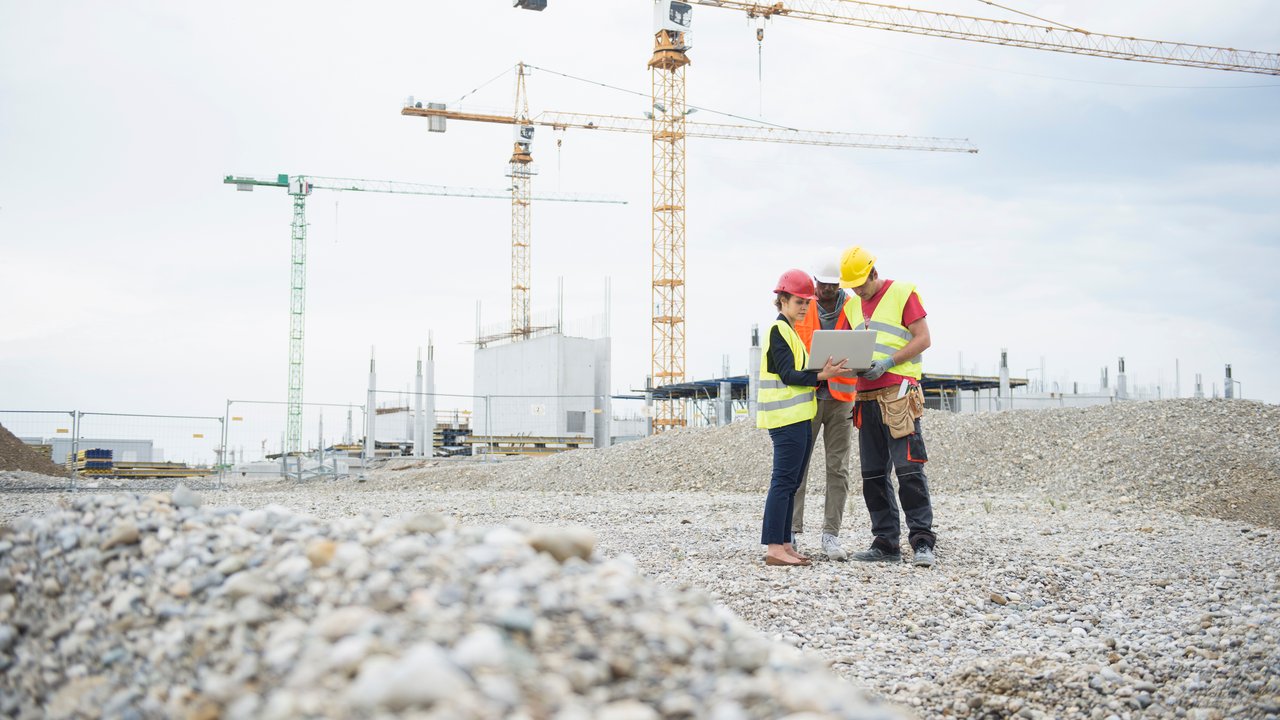 Two construction workers and a woman review plans on a laptop at a construction site with cranes in the background.