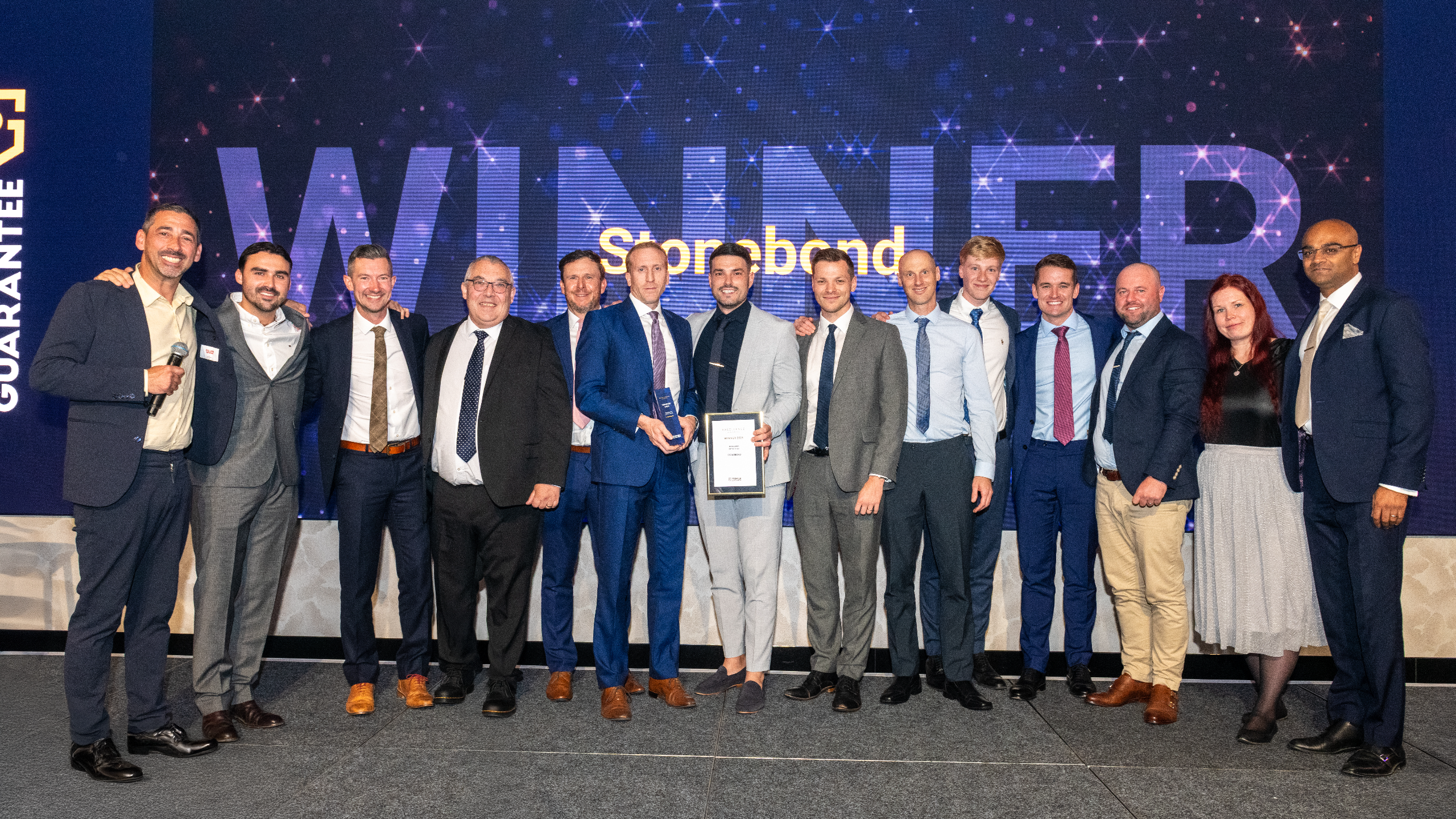 Excellence Awards 2025 11 A group of people celebrating an award on stage with a large "WINNER" sign in the background.
