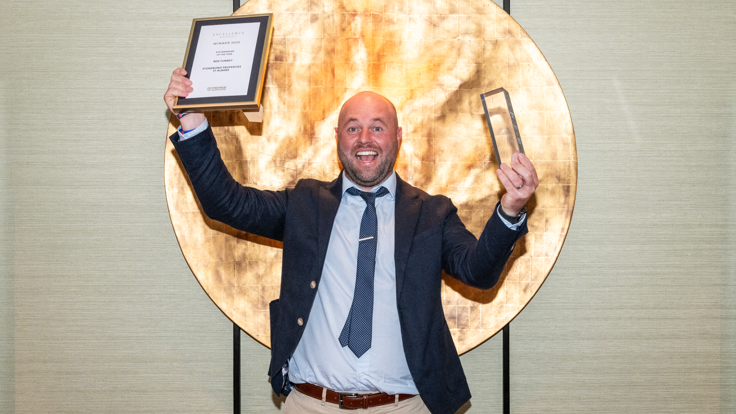 Excellence Awards 2025 13 A person in a suit holds a plaque and an award in front of a large circular backdrop.