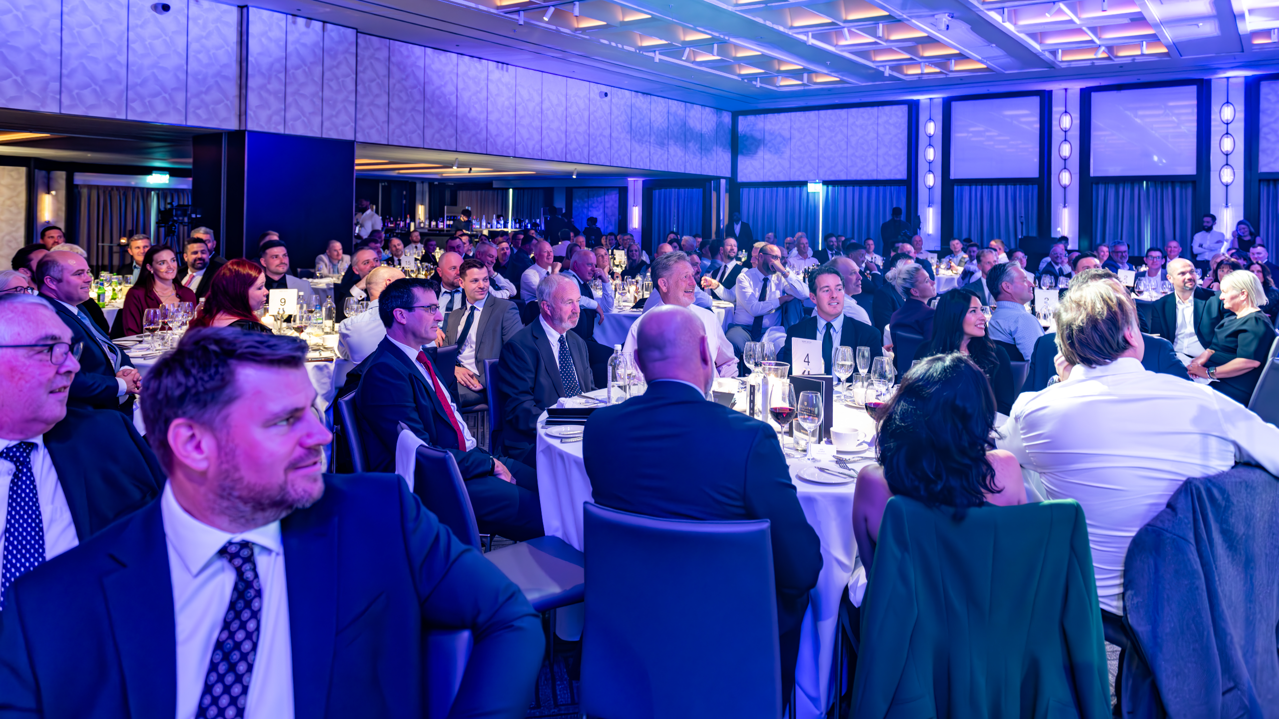 Excellence Awards 2025 3 A crowded banquet hall with guests seated at tables, enjoying a formal event with dim lighting and blue accents.