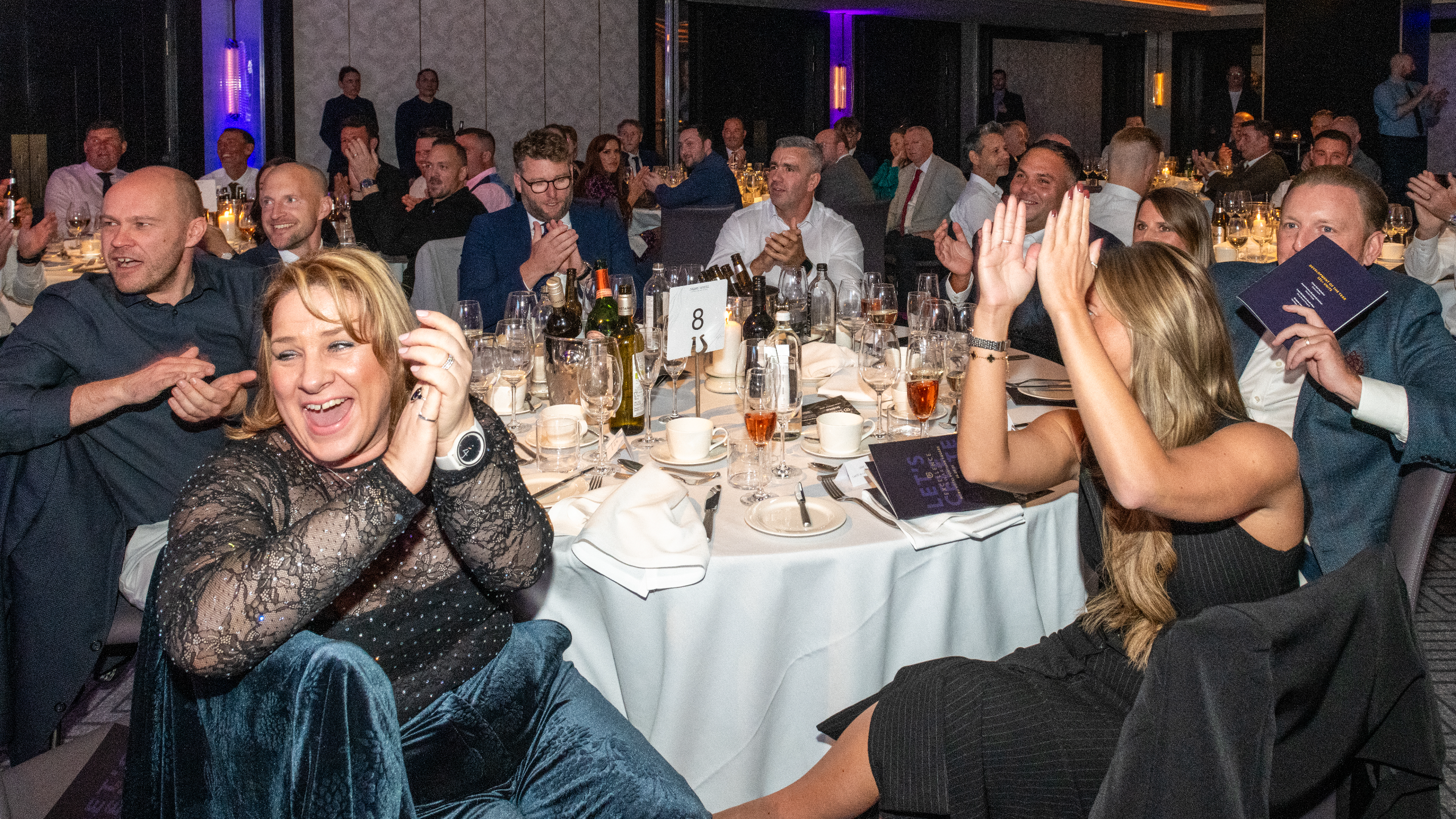Excellence Awards 2025 4 A group of people at a dining table, clapping and celebrating, with drinks and plates on the table.