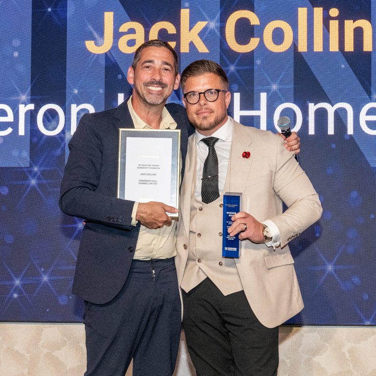 Two men pose together on stage, one holding an award certificate and the other holding a trophy.