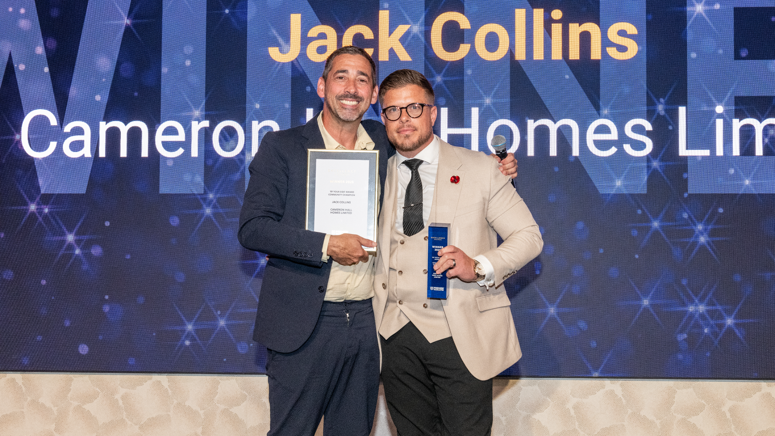 Excellence Awards 2025 6 Two men stand together on stage, one holding an award certificate and the other holding a trophy.