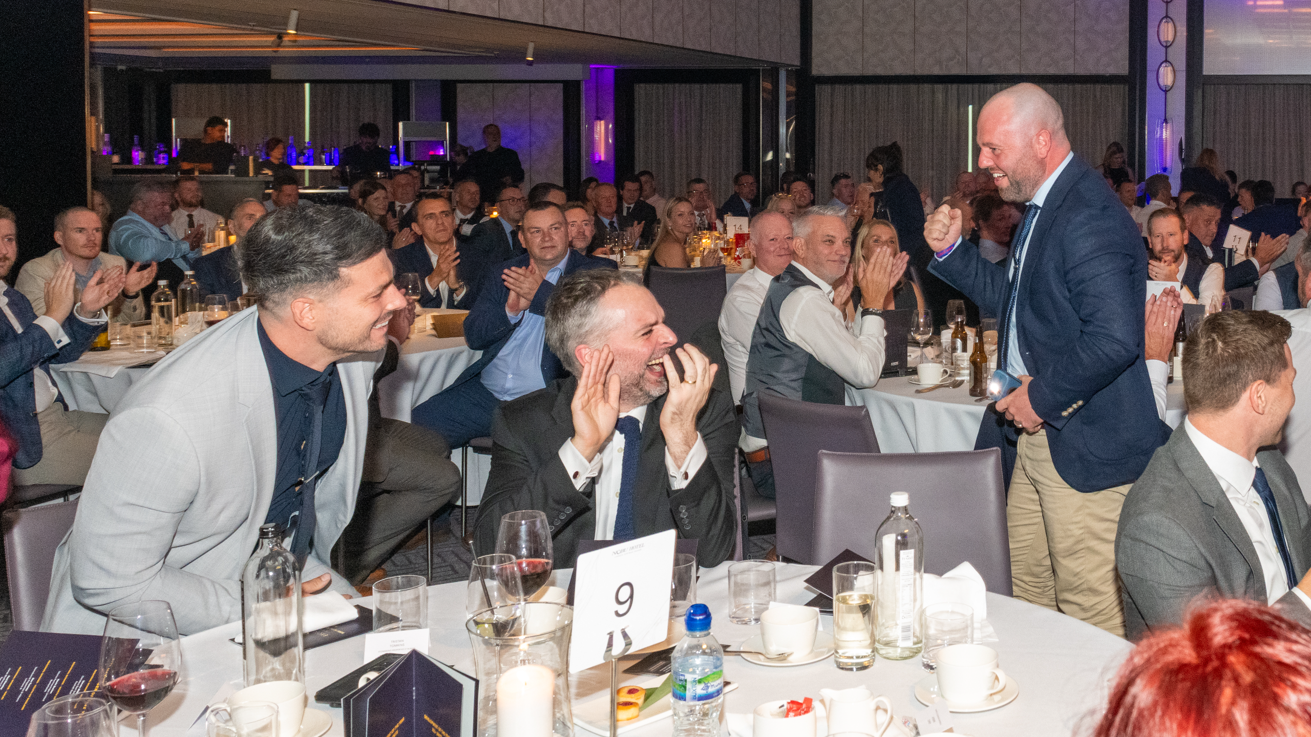 Excellence Awards 2025 8 A group of people at tables in a banquet setting, some clapping and celebrating, with drinks and plates visible.