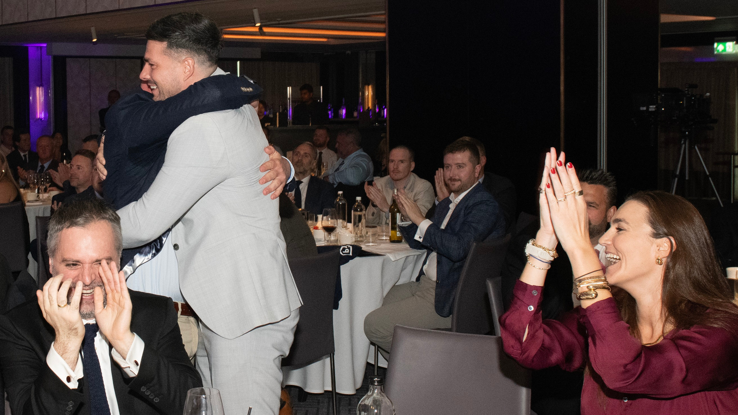 Two men embrace in celebration at a dining event, while others applaud and watch from their tables.