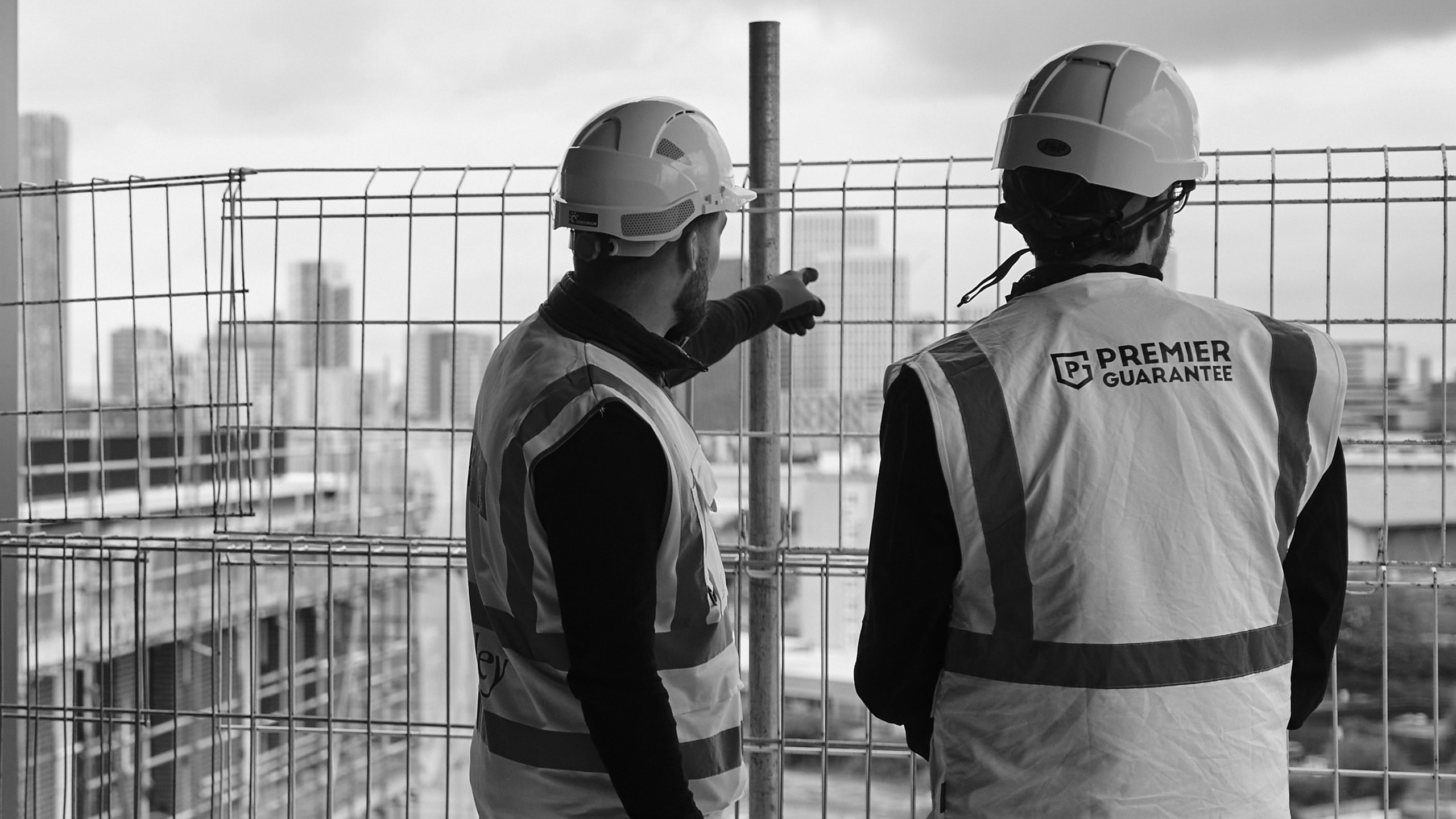 Two construction workers in safety vests and helmets observe a city view through a wire fence.
