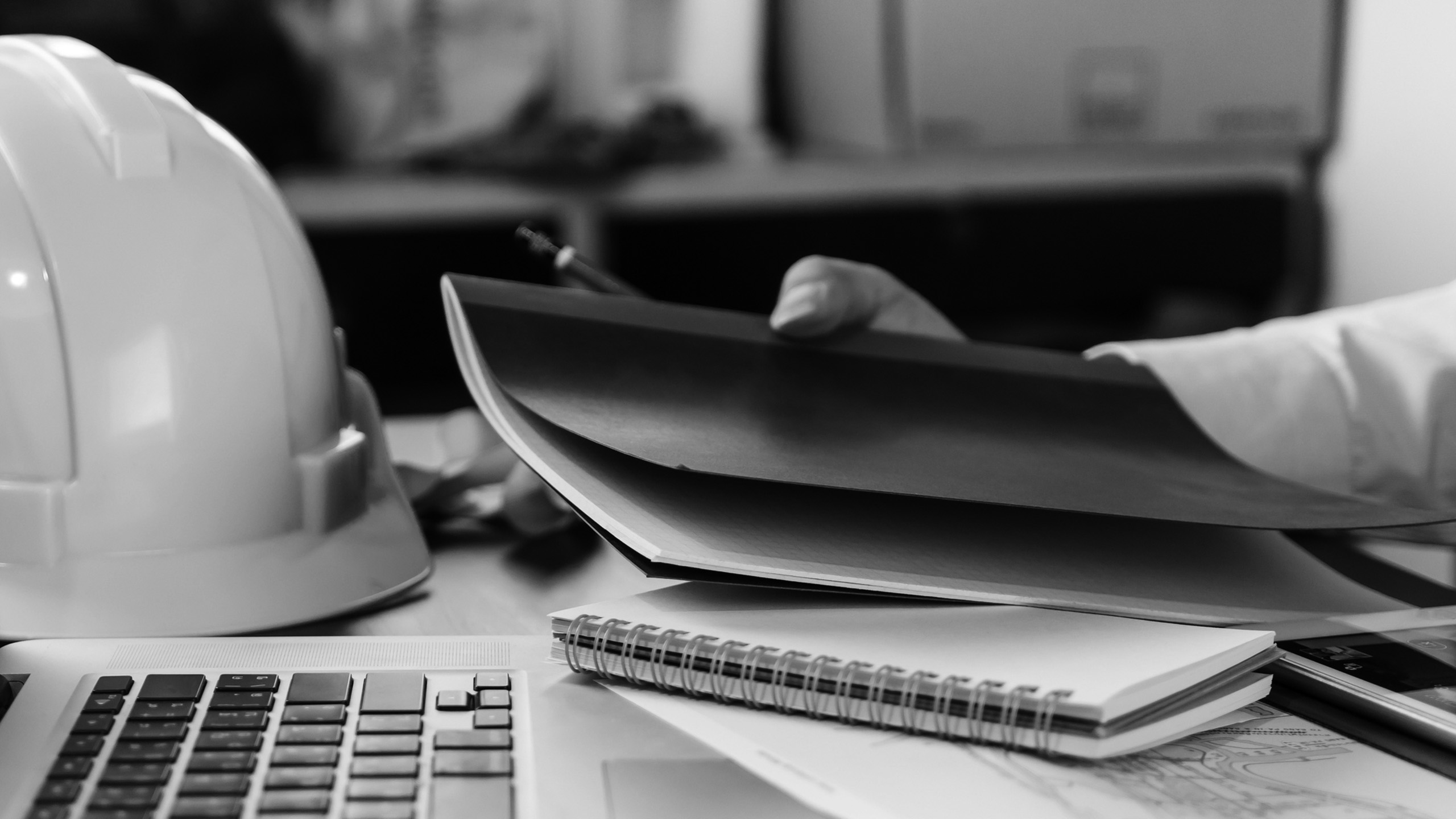 Brokers Close up of a man working on his laptop at his desk, with a hard hat on his desk in black and white