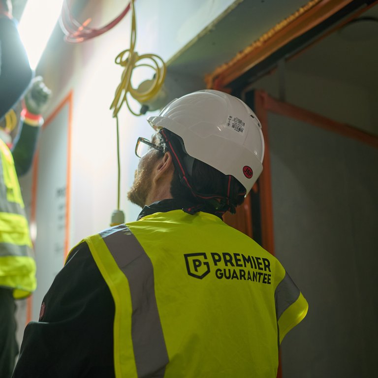 Two construction workers in safety vests and helmets, one working on a light fixture, the other observing.