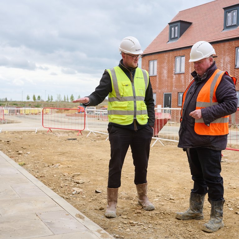 Two construction workers in safety gear discuss plans on a building site with houses in the background.