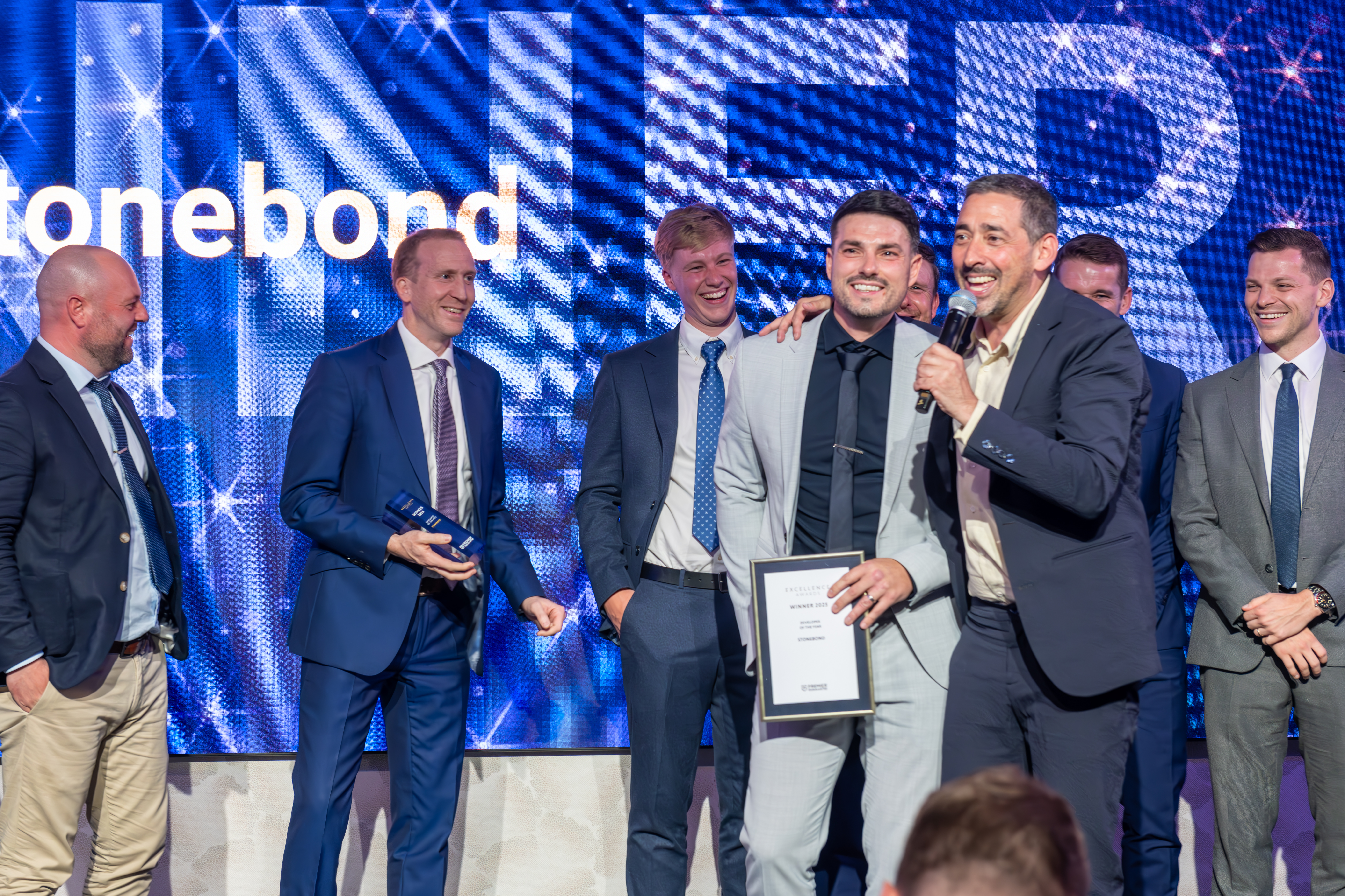 A group of men in formal attire standing on a stage with a "WINNER" and "stonebond" backdrop. One man is speaking into a microphone while another holds an award plaque.