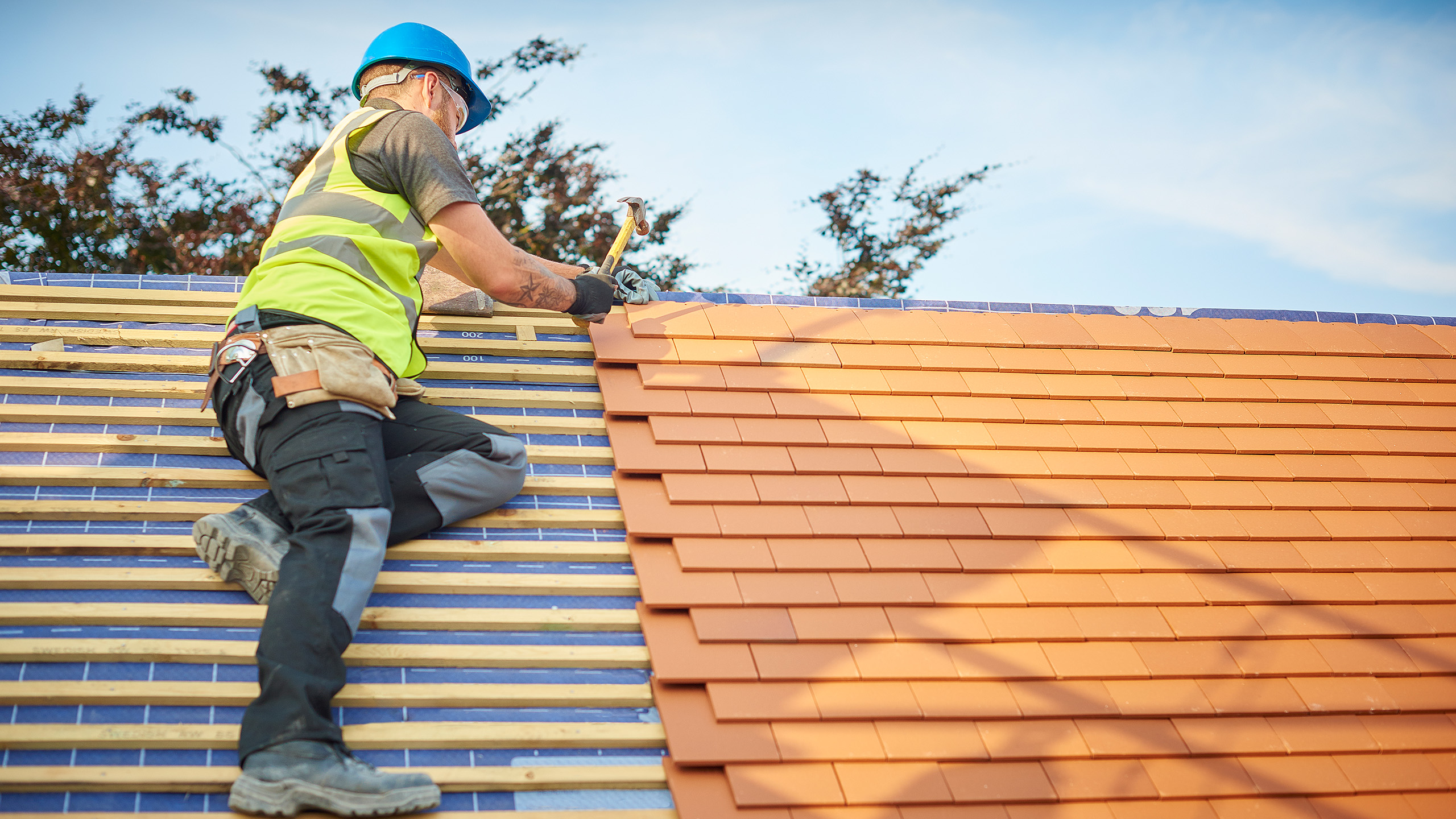A construction worker wearing a blue helmet and yellow safety vest installs red roof tiles on a house roof, kneeling and using a hammer, with trees and a clear sky in the background.