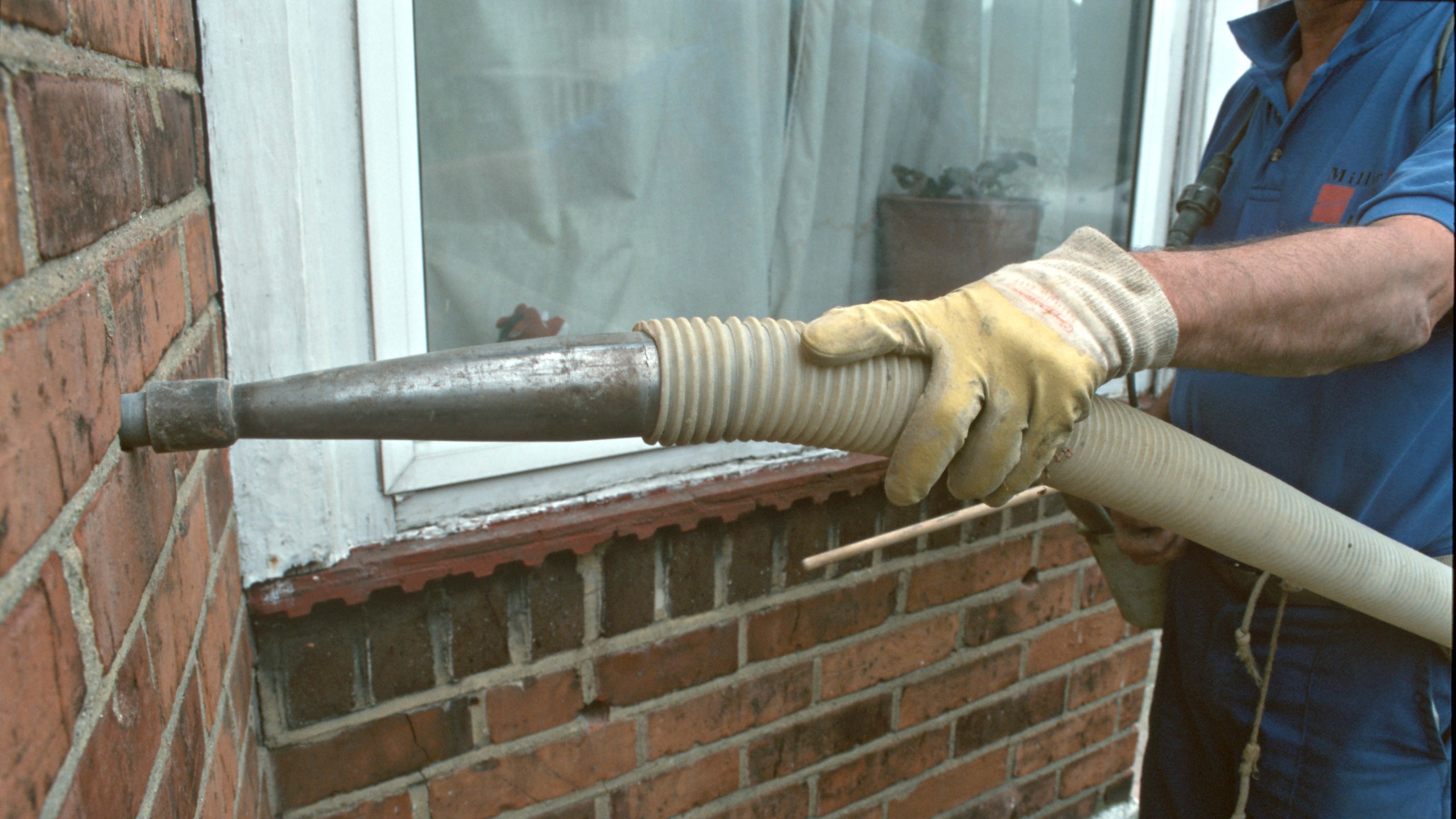 A person in gloves holds a hose attached to a wall, near a window, for maintenance work.