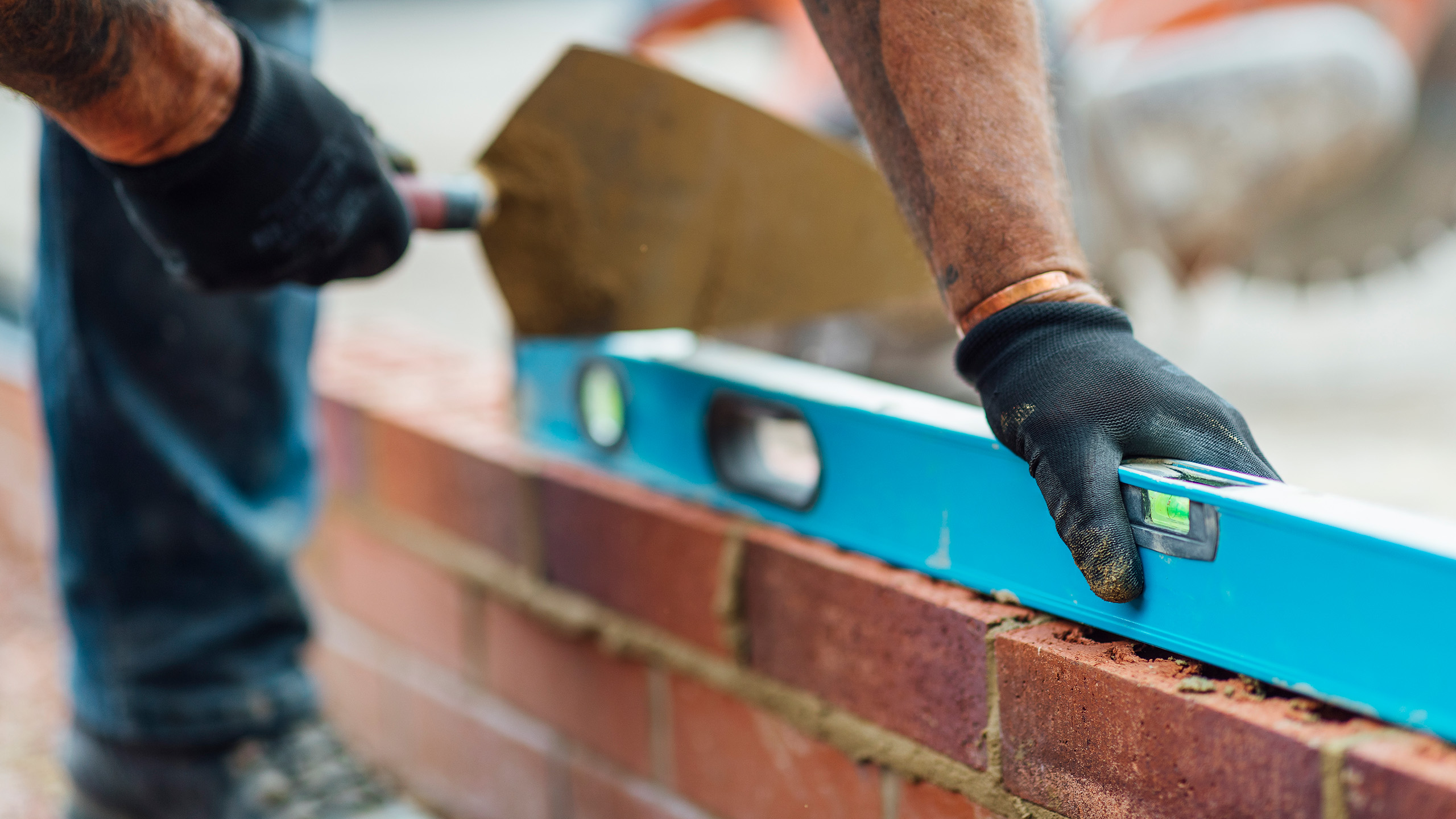 A close-up of a construction worker's hands using a blue spirit level to check brick alignment while laying a wall, with a trowel in the other hand and construction tools in the background.