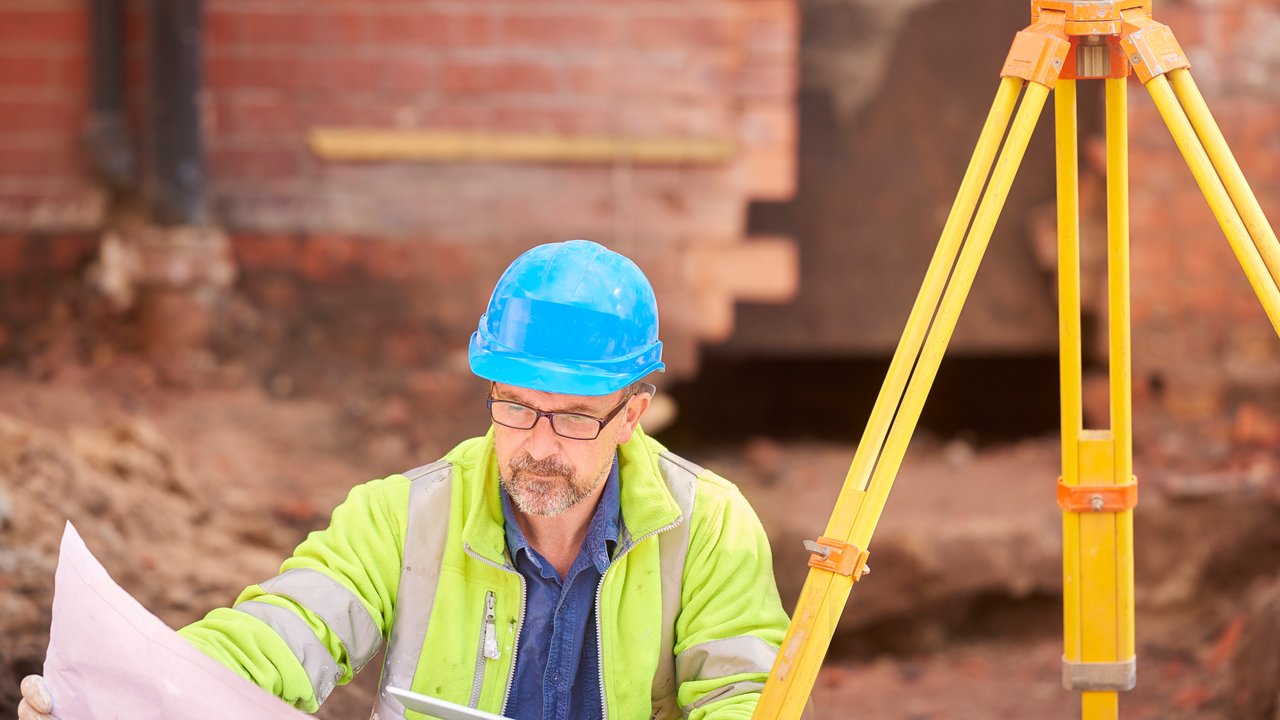 A construction site surveyor in a blue hard hat and high-visibility jacket examines a blueprint while pointing with a pencil. A yellow tripod-mounted surveying instrument stands nearby.