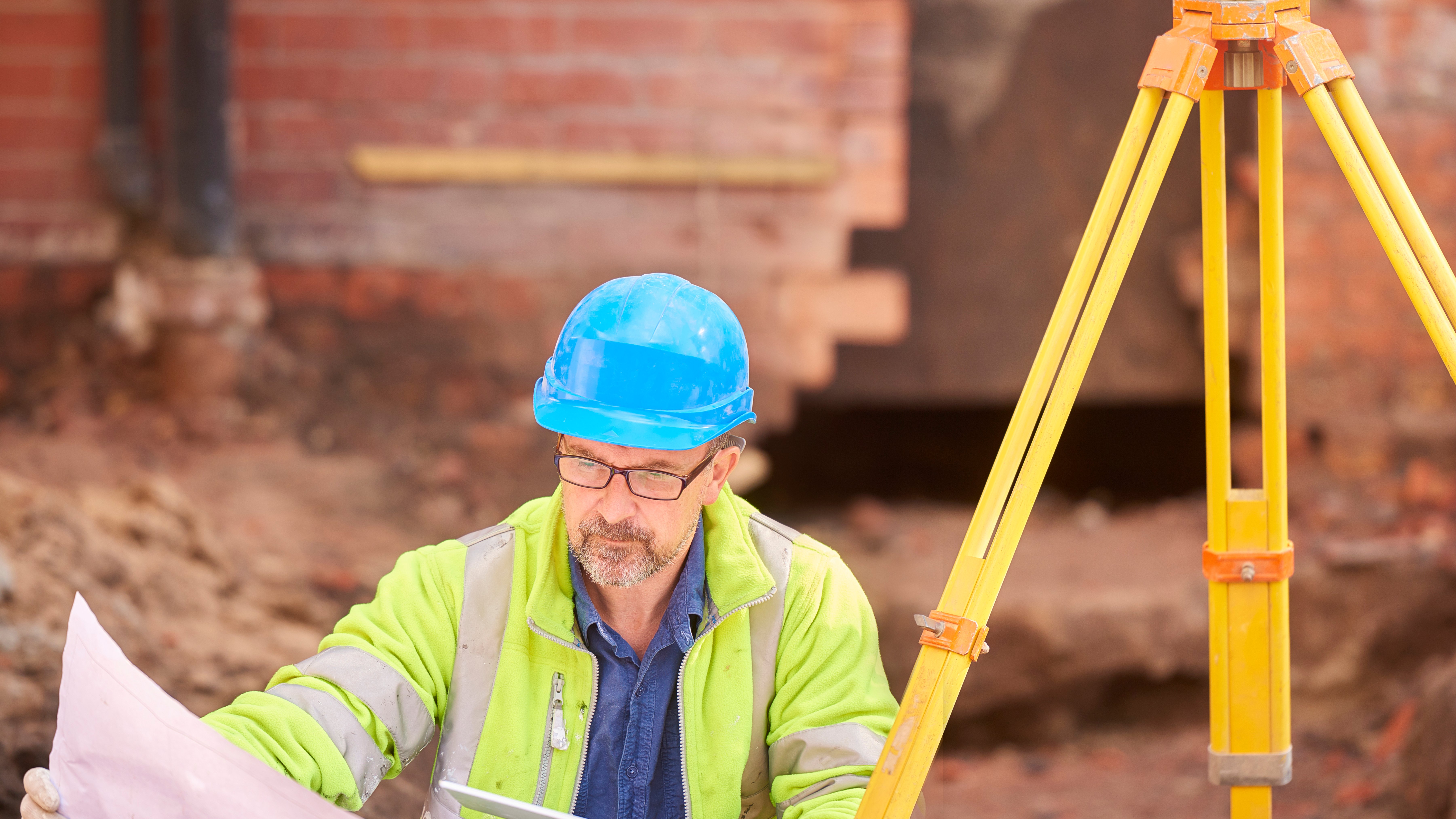 Construction Site Surveyor with Blueprint and Tripod Instrument A construction site surveyor in a blue hard hat and high-visibility jacket examines a blueprint while pointing with a pencil. A yellow tripod-mounted surveying instrument stands nearby.
