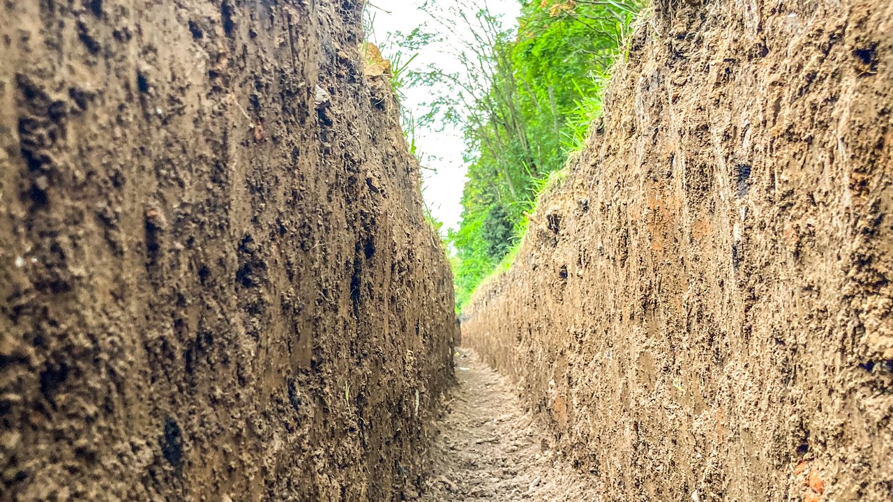 Excavated Soil Trench in Natural Landscape | Groundwork & Earthworks A narrow, deep trench dug into compacted soil with loose dirt at the bottom, surrounded by green foliage in an outdoor setting.