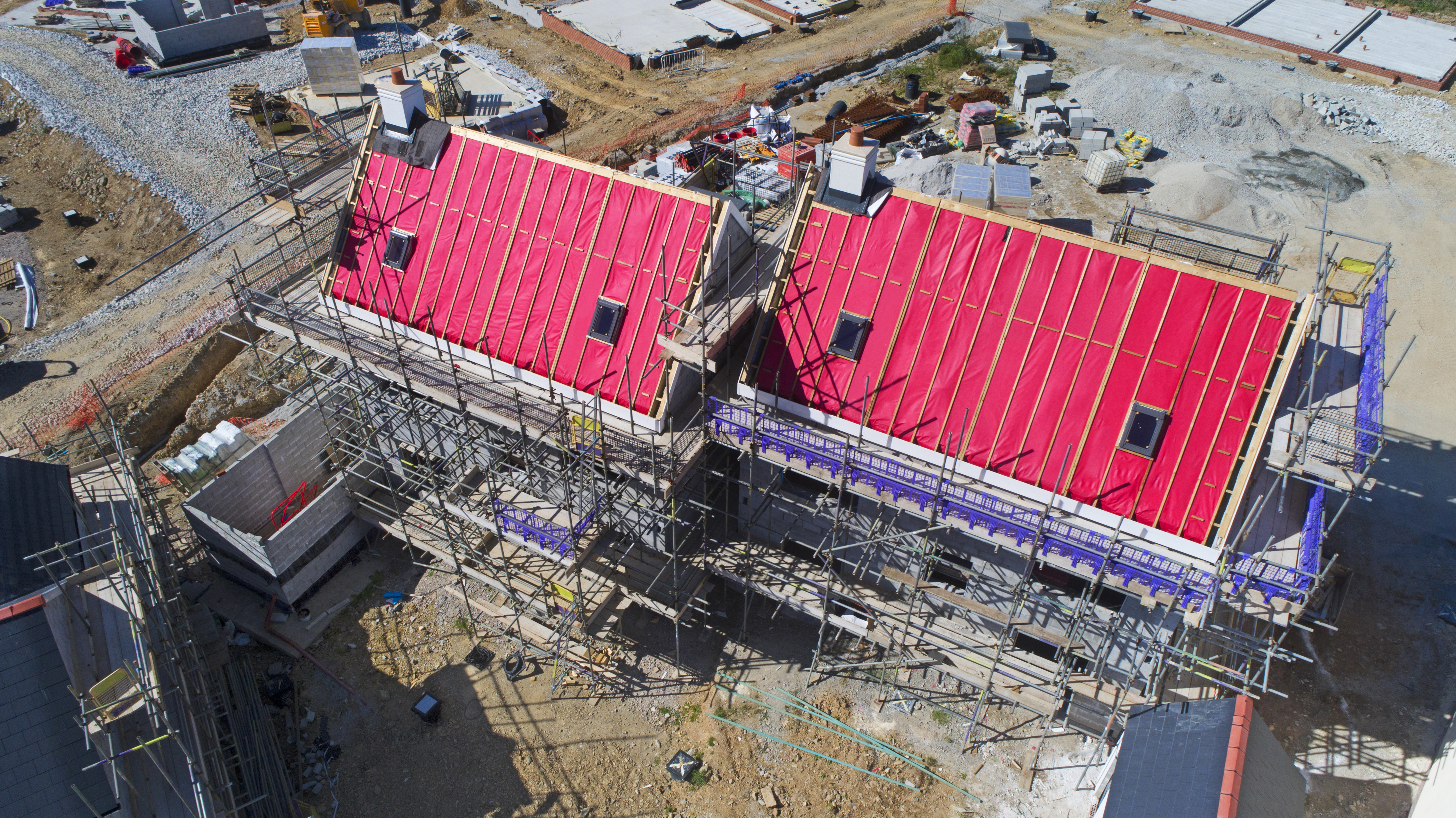 Aerial View of a Construction Site Aerial view of two buildings under construction with red roofing material and scaffolding surrounding them. The ground is covered with various construction materials and equipment.