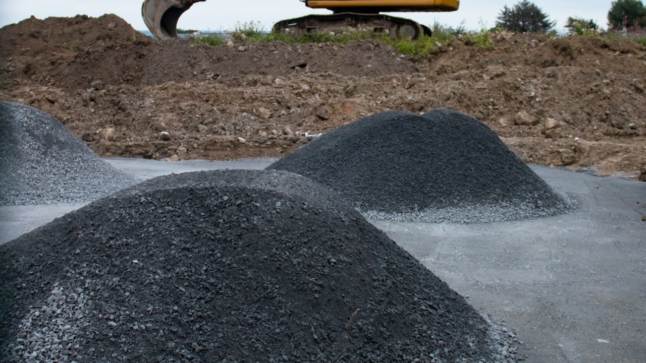 Pyrite contamination ellow excavator on a dirt mound with multiple conical gravel piles in the foreground at an active construction site under an overcast sky.