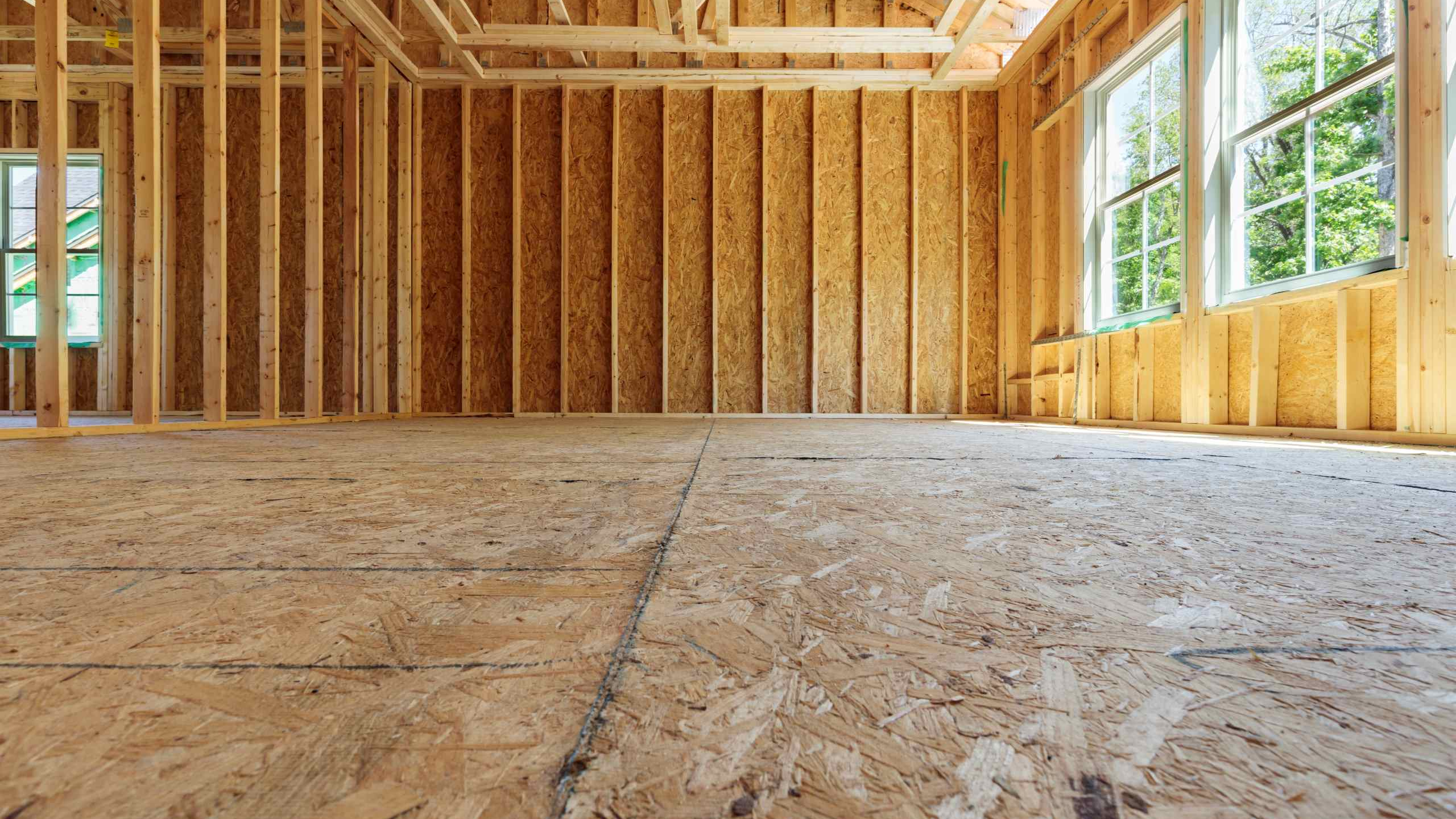 Interior view of a residential building under construction with exposed wooden framing, OSB subfloor, and large windows letting in natural light.