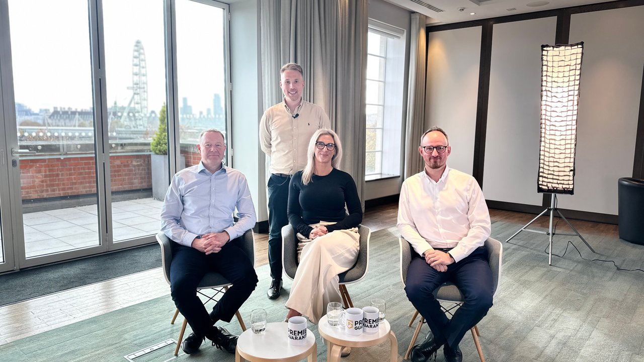 Four people seated indoors, with a city view and a light fixture in the background. Coffee cups on a table.