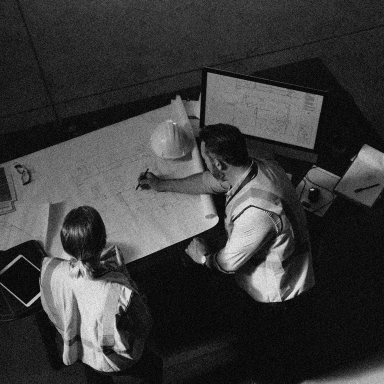 Two construction workers reviewing blueprints on a table, with a computer and safety helmets nearby.